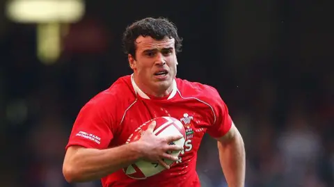 Getty Images Rugby player Jamie Roberts, with brown hair and wearing a red Wales shirt, clutches the rugby ball against his chest and runs