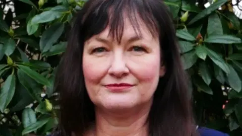 Head and shoulders image of Lynn Kelly, who has long dark hair and is standing in front of a rhododendron