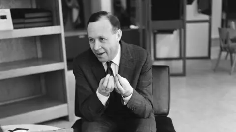 Getty Images A black and white image of a man in a with dark hair in a dark suit with a tie who is gesticulating with shelves and seats behind him