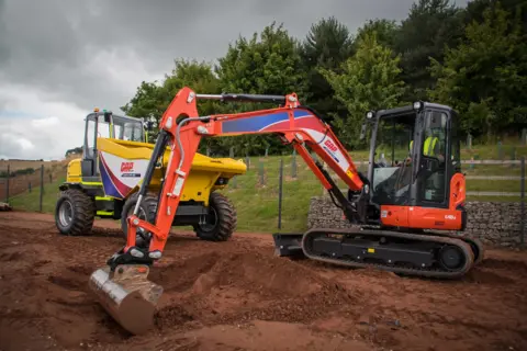 A yellow dumper trck and a red and black digger on an area of earth, next to a grassy bank and trees