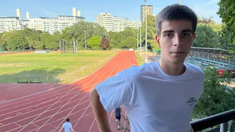 A man stands on a balcony overlooking an athletics track, wearing a white T-shirt.