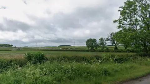 A grass field. Cars can be seen travelling on a road in the distance. The sky is overcast.