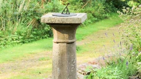 Julie Riley A sandstone sundial in a large garden with a pathway and flowers all around