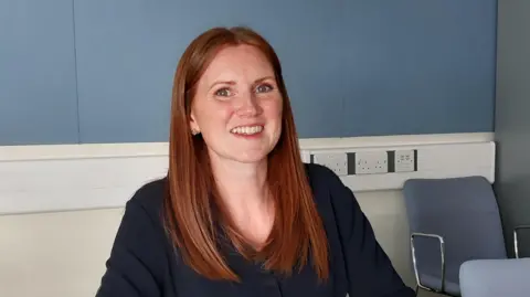 Essex Police A woman with long red hair sitting in an office and smiling at the camera. There are blue chairs and a blue wall behind her.