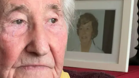 A close up of Mr Rayner looking away from the camera. He has brown eyes and white hair. There is a photo of a woman behind him in a white picture frame. She has brown hair.