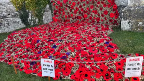 Guerrilla Gardeners A close-up of the poppy installation at ground level. Hundreds of knitted poppies are attached to the cargo net which is puddling at the bottom of the tower. A small chain has been placed around the artwork with signs saying - danger, please do not touch.