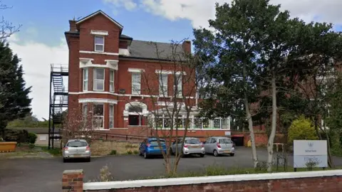 A three-story red brick building with a ramp entrance, conservatory and outdoor staircase seen set back from an off-road car park. A sign for the care home can be seen close to a low wall running along a pavement. 
