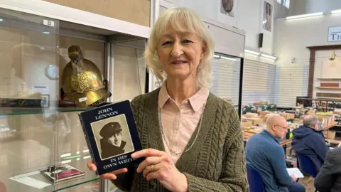 Lindsay holding a copy of the book. She is an older lady with white hair, and is wearing a green cardigan. Behind her is a shelving unit with other items for sale