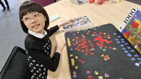 Family photo Ariel sitting at a table in front of a puzzle piece. She is looking away from teh camera.
