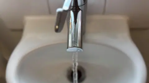 A shiny silver tap expels a strong flow of water into a white sink. The handle of the tap is over the middle of the outlet, and the water is pouring out directly over the sinkhole. The basin is attached to a white tiled wall.