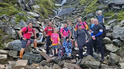 Pete Haslam A group of 11 people posing at the bottom of Ben Nevis, the terrain is rocky. All looking at the camera and smiling, wearing climbing gear and equipment. Ted is also there in his wheelchair.
