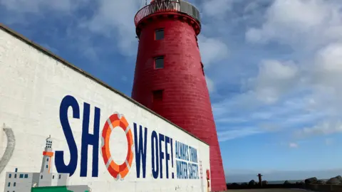 A white wall with a mural leads up to a red lighthouse. The sea is behind the lighthouse, along with rocks. The sky is blue with some white clouds. 