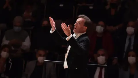 James Ross in formal concert attire stands on a podium with both hands raised during a performance in a concert hall. The conductor is illuminated by a spotlight in the foreground, while rows of audience members sit in the dimly lit background. Many audience members are wearing face masks. 