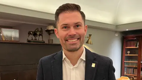 Darren Rozier/BBC Tom Slegg, the manager of The Angel Hotel. He is looking directly at the camera and smiling. He is wearing a white shirt with a blazer over the top. He is standing inside the hotel with a bookcase behind him. 