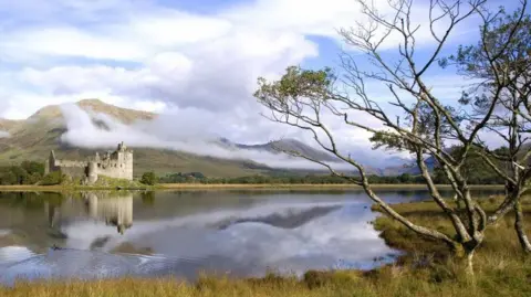 Getty Images A view of Loch Awe looking towards Kilchurn Castle. The water is clear and is reflecting the sky, which is blue. There is a tree in the foreground on some long green grass.