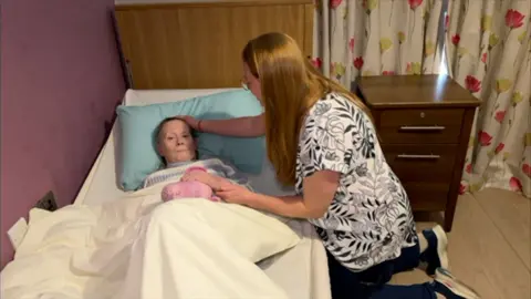 A woman lying in a care home bed with her daughter, a woman with a black and white patterned top and long red hair, by her side