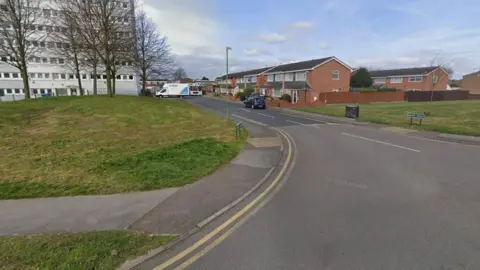 Google A Google street view image of the entrance to a residential street. A green space is on the left with several trees on it. A tower block is visible behind them. On the right are a number of homes. Several vehicles are parked on the road.