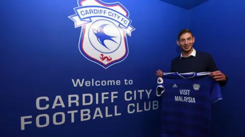 Getty Images A young man with brown hair, wearing a white shirt and dark jumper. He is holding a blue Cardiff City football shirt and is stood in front of a blue, Cardiff City branded wall which reads "Welcome to Cardiff City Football Club". He is smiling and looking at the camera.