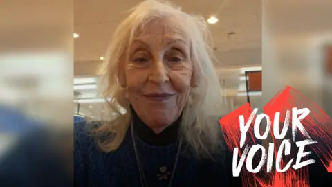 A woman with grey hair in a brown jumper with a Your Voice logo next to her