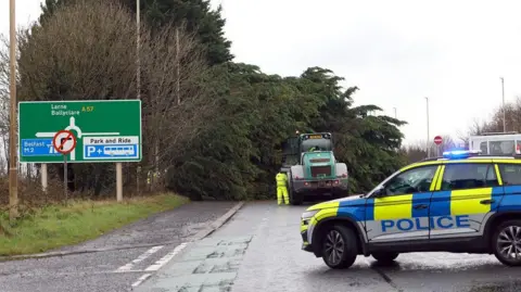 Pacemaker Police car and tractor on road with large fallen tree blocking path, man in hi viz visible in distance working on tree