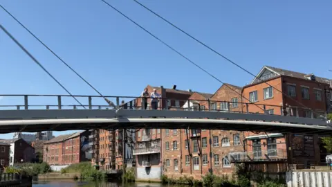 Leeds Museums and Galleries A bridge crossing a river. Two people are stood on it and looking across the water.