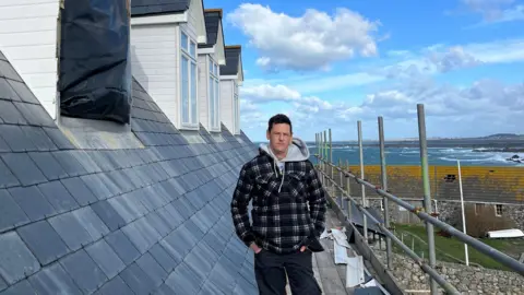 Steve Sarre stood on scaffolding on the house in lihou. It shows damage to windows.