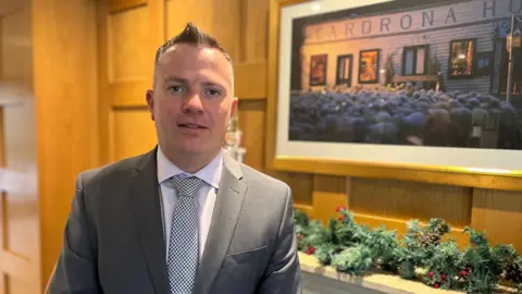 A man with short brown hair, wearing a grey suit, white shirt and light blue tie, looks at the camera, with a decorated fire place in the background