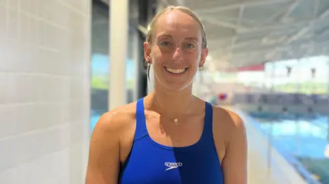 Hannah is in a blue swimsuit stands beside an indoor swimming pool. The pool, lane markers and bright leisure centre interior are visible in the background, with the water and pool area softly out of focus.