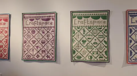Craftspace Four giant quilts hang in a gallery. Each is a patchwork depicting everyday objects and each have the name Craftwpace at the top in patchwork lettering. 