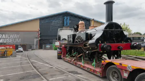 The Board of Trustees of the Science Museum Group A steam engine is on the back of a low-loader vehicle which is being driven away from the camera toward a large building with the words Railway Museum in large white letters on the left hand side of the building.
