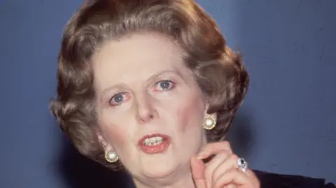 Archive photograph of Margaret Thatcher taken in 1984. She has dark-blonde hair and blue eyes, is wearing pearl-stud earrings and is holding her thumb and forefinger together as she makes a speech. On her third finger is a jewelled ring.