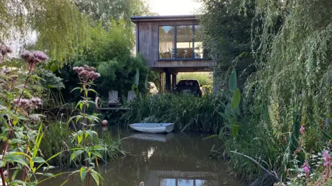 Iain Davies A timber house on stills above a lake. There's greenery all around and the house has huge windows to overlook the views. There's a little boat on the water.