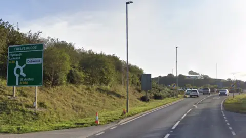 A road sign showing a roundabout leading to the A30 to Plymouth and Liskeard to the left, Dobwalls to the right and the A390 to St Austell further right. The sign sits on a grassy bank along the side of a single carriageway. There are cars gathering at the roundabout in the distance.