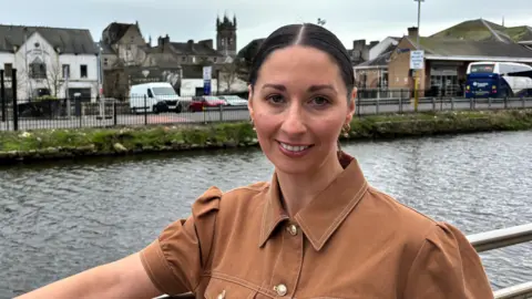 Rachel Duffy stands in front of Newry Canal. She is wearing a tan coloured shirt and has her dark hair pulled back from her face,
She is smiling