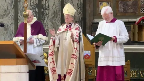 John Sherrington wears an ornately decorated cream and gold mitre and a white and red robe. He holds his hands out as he speaks in a cathedral. Two men in maroon and white robes stand either side of him,