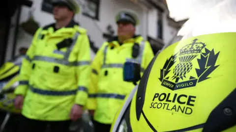 Two police officers wearing fluorescent yellow jackets are blurred in the background of the image. The front of a police motorcycle is visible in the foreground with the Police Scotland logo.