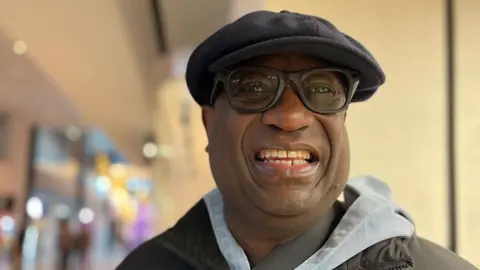 Emma Baugh/BBC Bruce Choto standing inside Queensgate Shopping Centre. He is smiling broadly and wearing a peaked grey cap, heavy black-framed glasses and a black jacket with a light grey collar. 