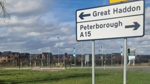 A housing estate in the distance with what look like newly built houses. Tall corporate flags are dotted around and there is a huge billboard in front of the estate. In the foreground of the photo is a white road sign. On top is an arrow pointing left next to the words "Great Haddon". Below is an arrow pointing right with the words "Peterborough" and A15.