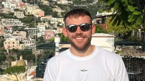 Sam Childerley A man is pictured smiling at the camera. He has short dark hair and a beard and is wearing sunglasses and a white T-shirt. 