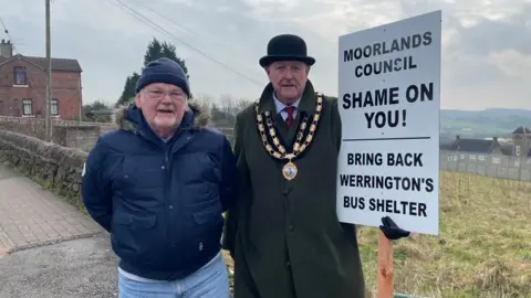 Two men standing on the pavement with field behind them. They are high up as fields can be seen in the background. The man on the left is wearing a navy puffer coat and navy hat,. The man on the right is wearing a long, dark green coat and a gold medallion, like a mayor would wear. He is holding a placard which reads "Moorlands council, shame on you. Bring back Werrington's bus shelter" in black writing. 