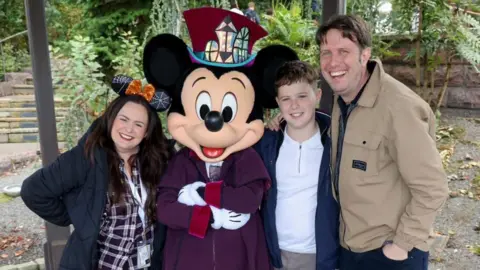 Fleming family Lisa Fleming has long dark hair. She is wearing Minnie Mouse ears, a black puffer jacket and a burgundy and white checked shirt. To her right is a person in a Mickey Mouse costume. To his right, Lisa's son is a dark-haired teenager in white T-shirt and blue jacket. To his right, his dad and Lisa's husband has brown hair and is wearing a light brown jacket and dark jeans. They are all laughing.