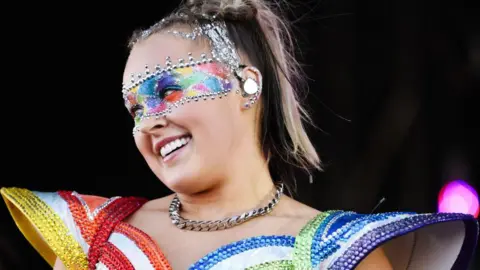 Getty Images JoJo Siwa smiling portion    performing connected  signifier    wearing a rainbow-coloured outfit with ample  enarthrosis  pads. She's wearing multi-coloured oculus  make-up which is framed by metallic  jewels and has a matching necklace astir   her neck.