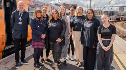 A group of eight women and one man stand on a railway platform next to an orange train. They smile at the camera. Several of them wear a black charity shirt which says 'I get it'.
