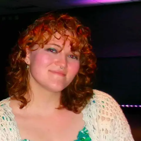 Hafwen Davies A portrait photograph taken indoors in a dark entertainment venue. One person is positioned in the centre of the frame, facing the camera from slightly above waist height. The person has shoulder-length curly red hair and is wearing a white open-knit cardigan over a green-and-white top decorated with a leafy botanical pattern. Small facial piercings are visible on the nose and ears. The lighting is low, with purple and pink light cast across the ceiling and faintly visible in the background. Most of the background is dark, making the person stand out against the surroundings. 