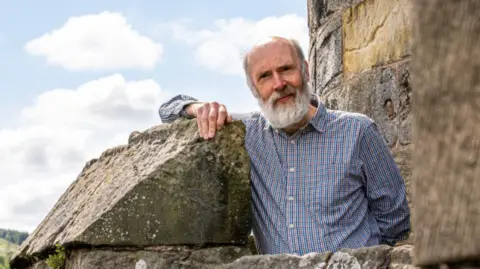 Falkland Estate Ninian Stuart, who has a white beard, leans on a large wall outdoors, with a cloudy sky in the background.