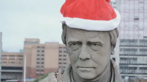 The grey face of a statue with a Santa hat on its head