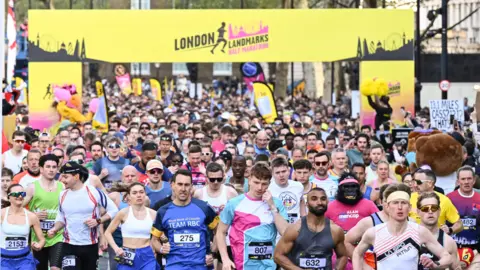 PA Media A massive crowd of runners wearing colorful athletic gear and race bibs stream through the starting gate of the London Landmarks Half Marathon. The large yellow overhead banner features the event logo and silhouettes of the London skyline.