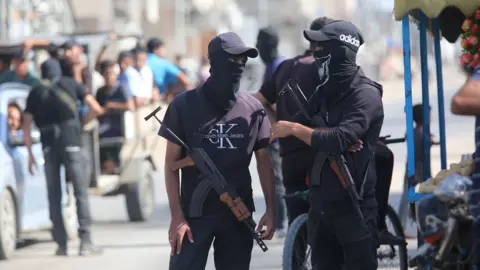 Anadolu via Getty Images Two security members wearing hats and balaclavas hold assault rifles while they converse as they stand on a street corner in Deir al-Balah on 10 October