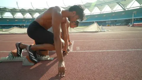 Abhay and Simran Crouch on the initial line to practice the beginning of a race. He is in the foreground with black shorts and a bare torso, and he is behind him in white.