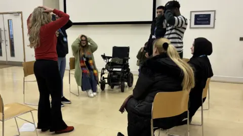 Gemma Dillon/BBC Image shows a class with 5 pupils either sat or stood in a circle. The teacher is wearing a red jumper and has her hands on her head as they are all playing a language game together